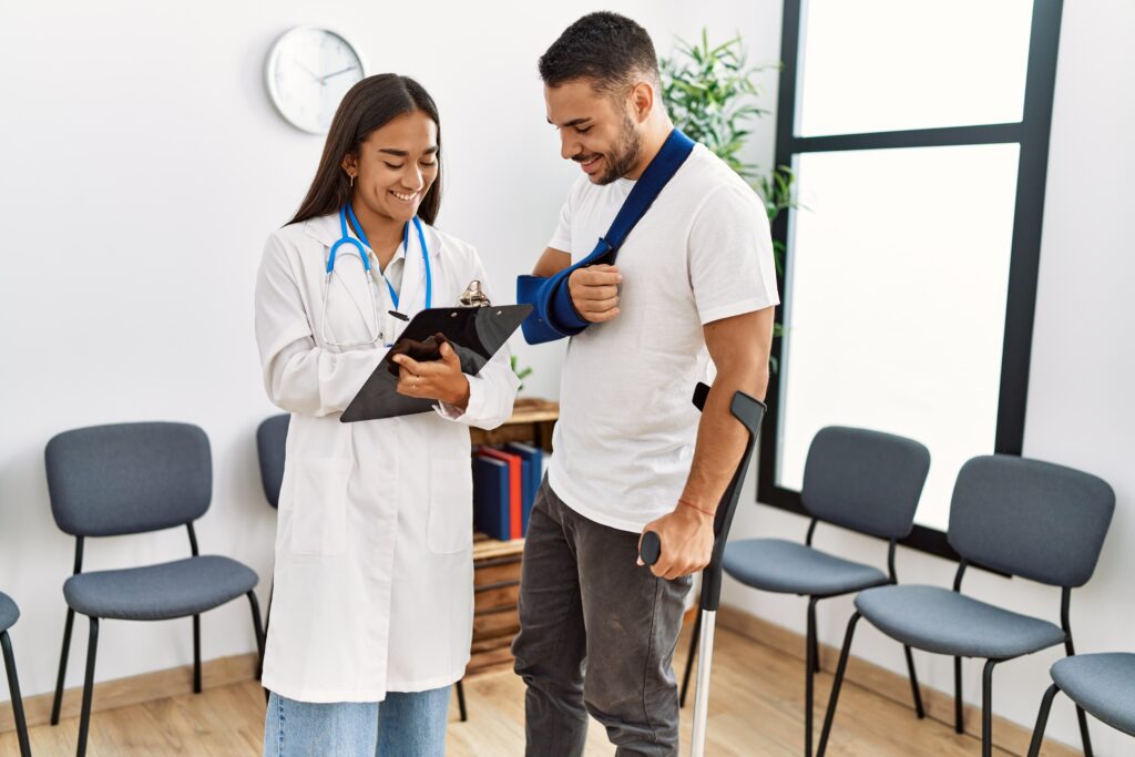 A doctor with long brown hair talking to a patient with a sling and crutches in a medical waiting room