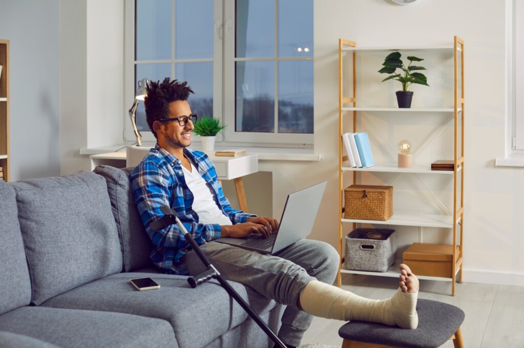 a person with short coily dark brown hair wearing a blue plaid shirt and a leg cast and sitting on their couch with their laptop and crutches nearby