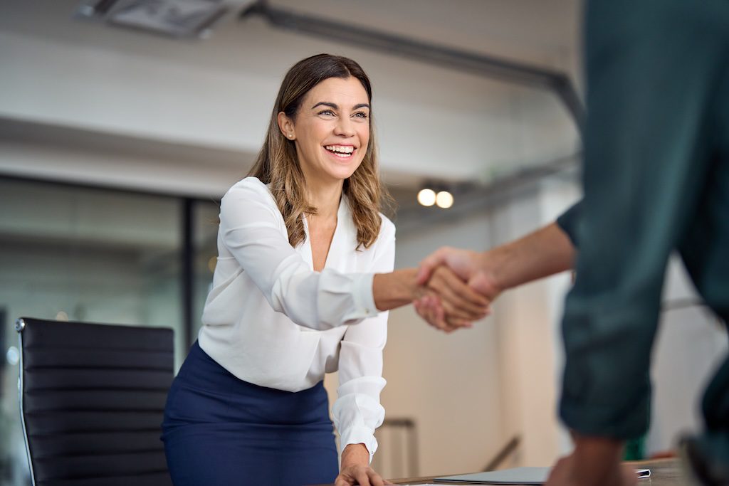 a person with long dirty blonde hair wearing business attire and smiling while shaking hands with someone not in the frame