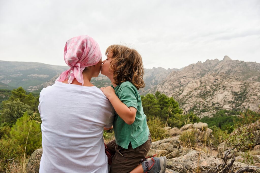 back view of a person wearing a pink head scarf and white shirt being kissed on the cheek by a young child while sitting outside on a hiking trail