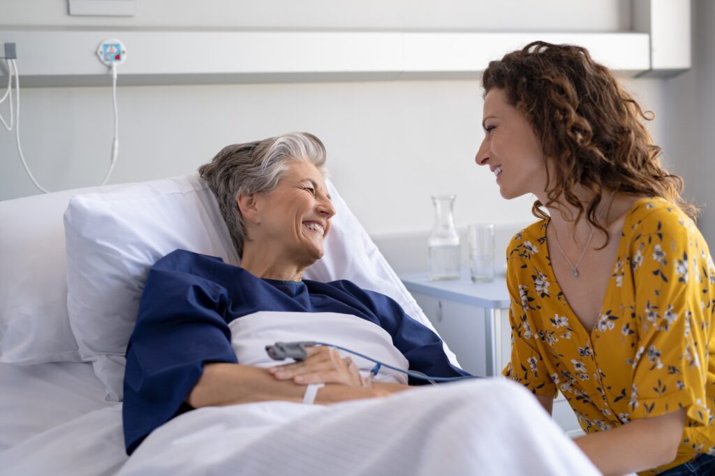 a person with short grey hair lying in a hospital bed and smiling at a person with long curly red hair wearing a yellow flowered blouse